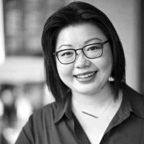 A woman with medium-length hair stands against a plain white wall, smiling softly. She wears a collared jacket over a blouse, and the grayscale image creates a clean, approachable impression.