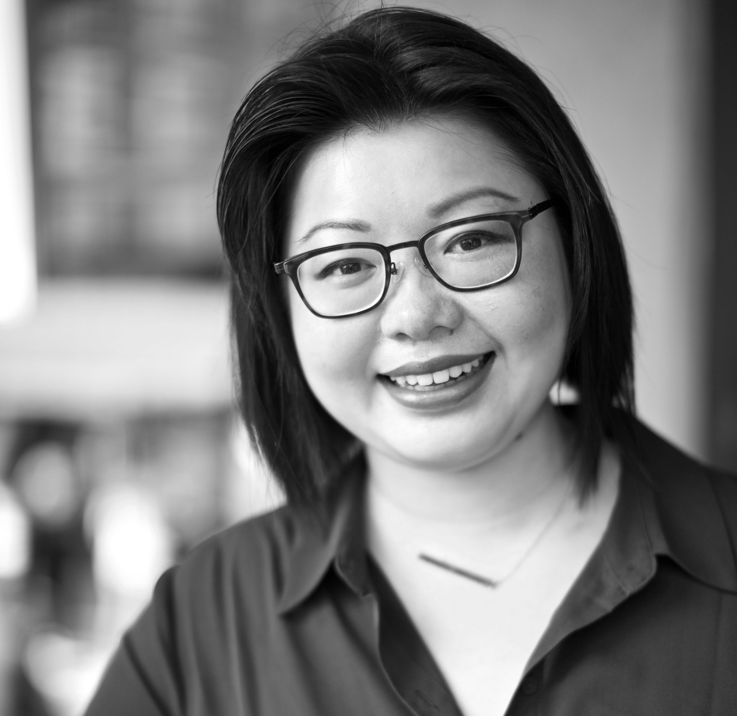 A woman with medium-length hair stands against a plain white wall, smiling softly. She wears a collared jacket over a blouse, and the grayscale image creates a clean, approachable impression.
