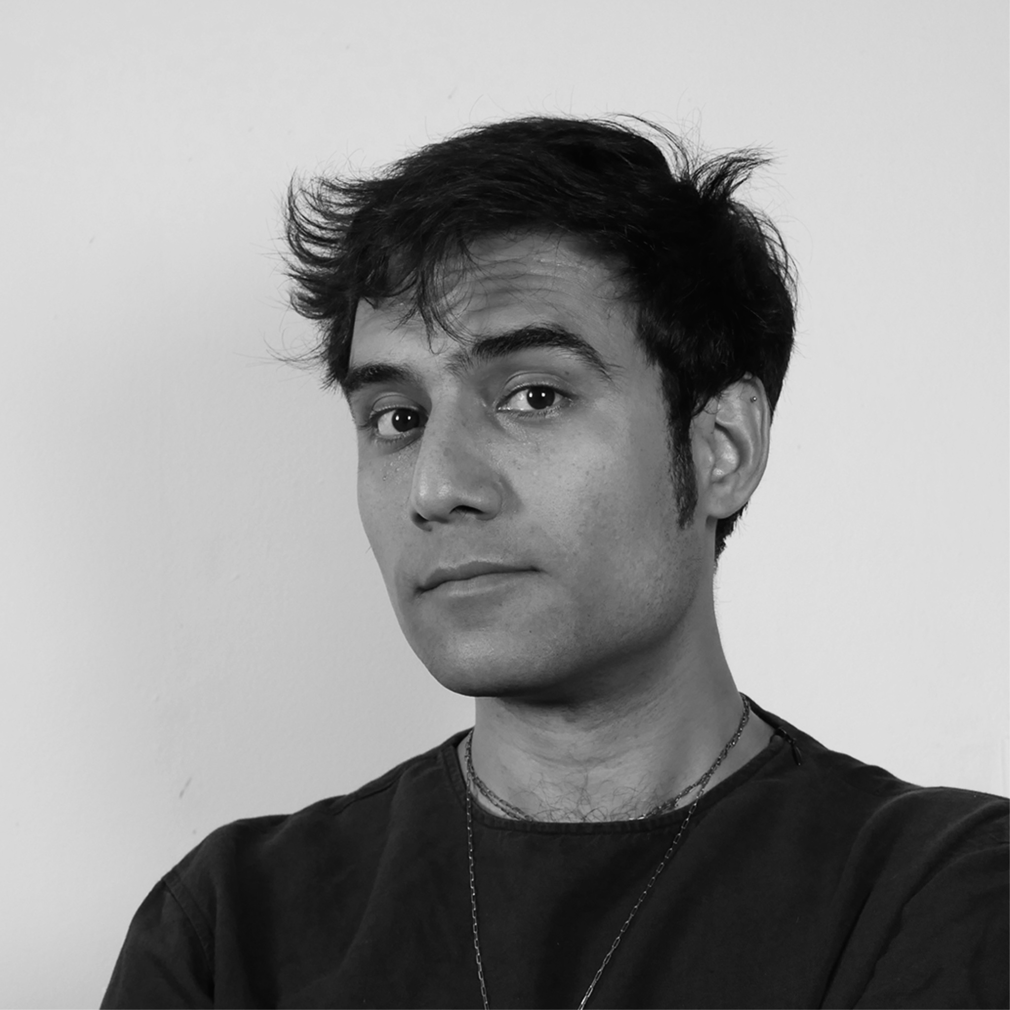 A young man with dark, tousled hair looks slightly to the side with a raised eyebrow. He wears a dark shirt and a chain necklace. The background is plain and light-colored. The portrait is in black and white.