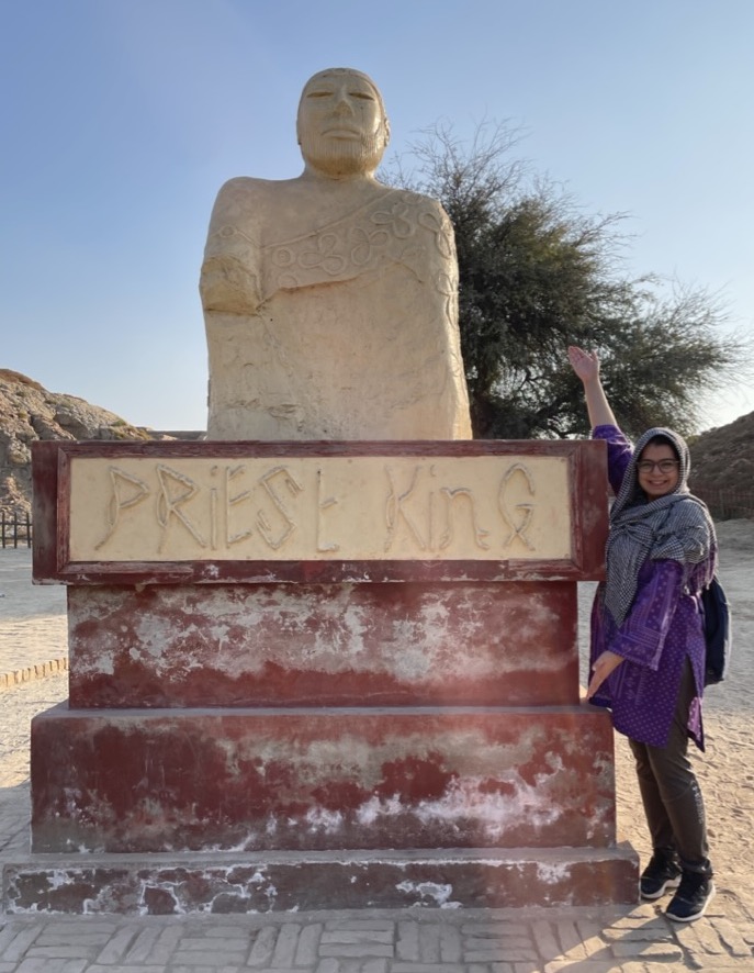 A woman in a headscarf looking at the camera stands next to a sculpture in an arid landscape. The base of the sculpture reads, "Priest King."