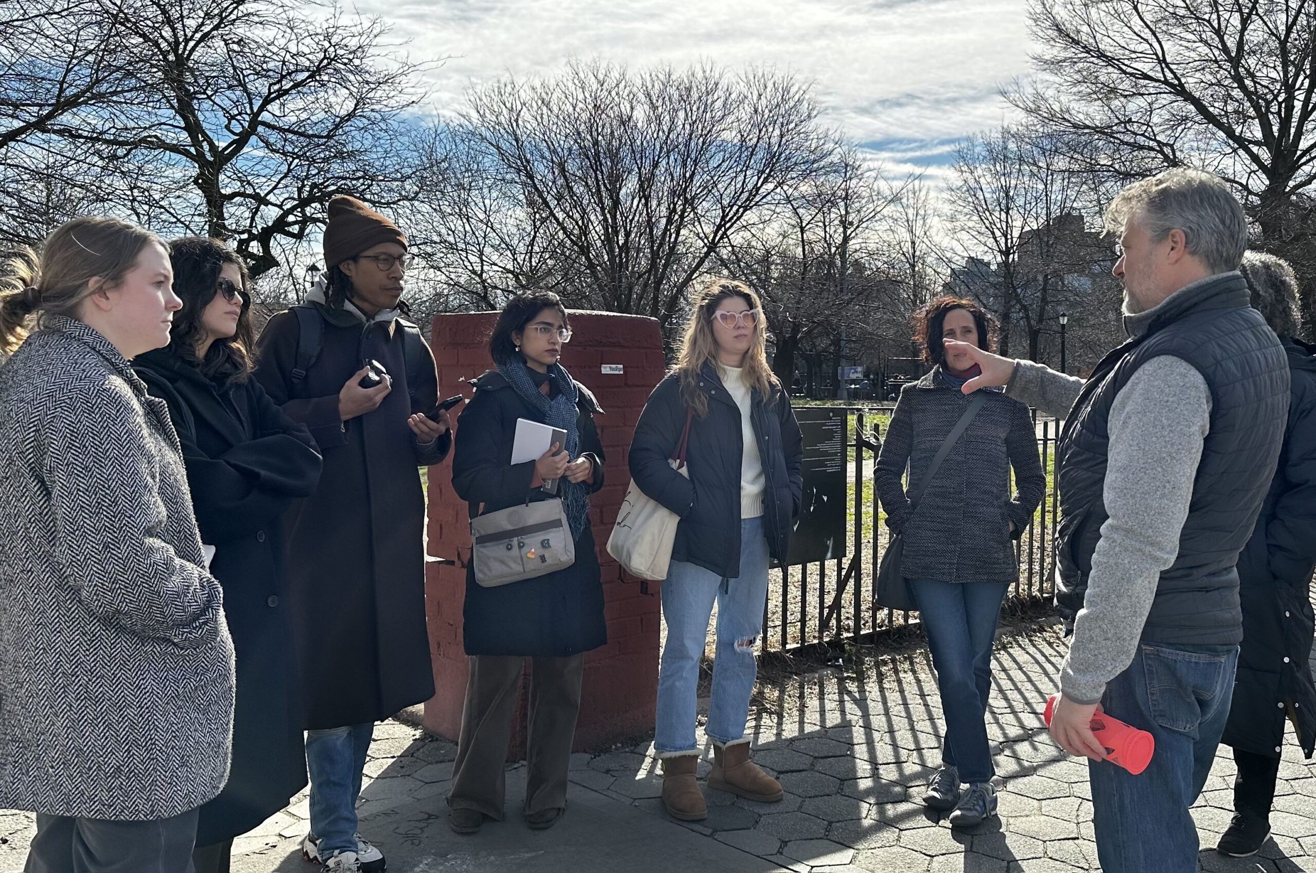 A group of students stand outside by a park attentively listening to someone speaking.