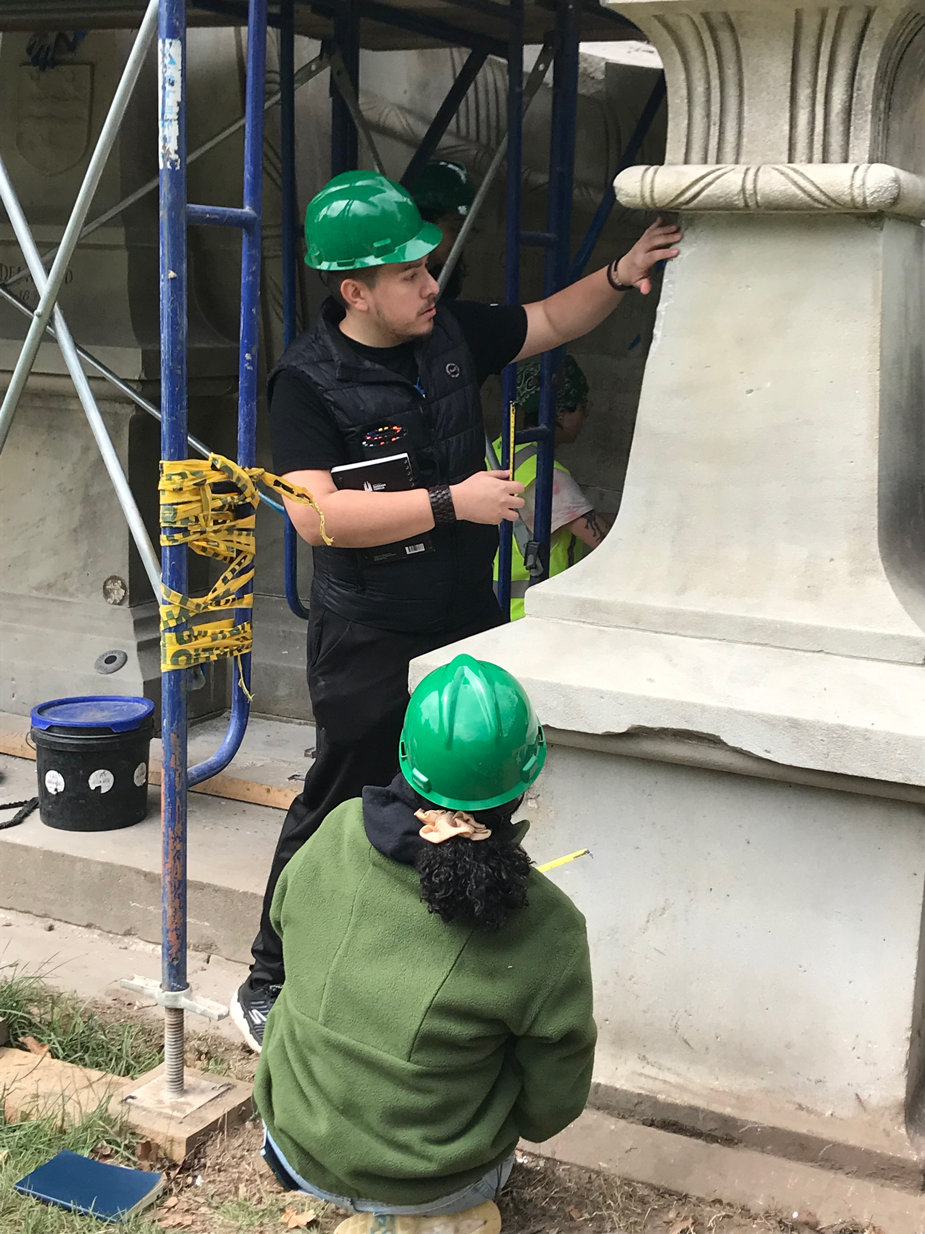 Two students work on masonry in Green-Wood cemetery. They are wearing hardhats and there is scaffolding behind them.
