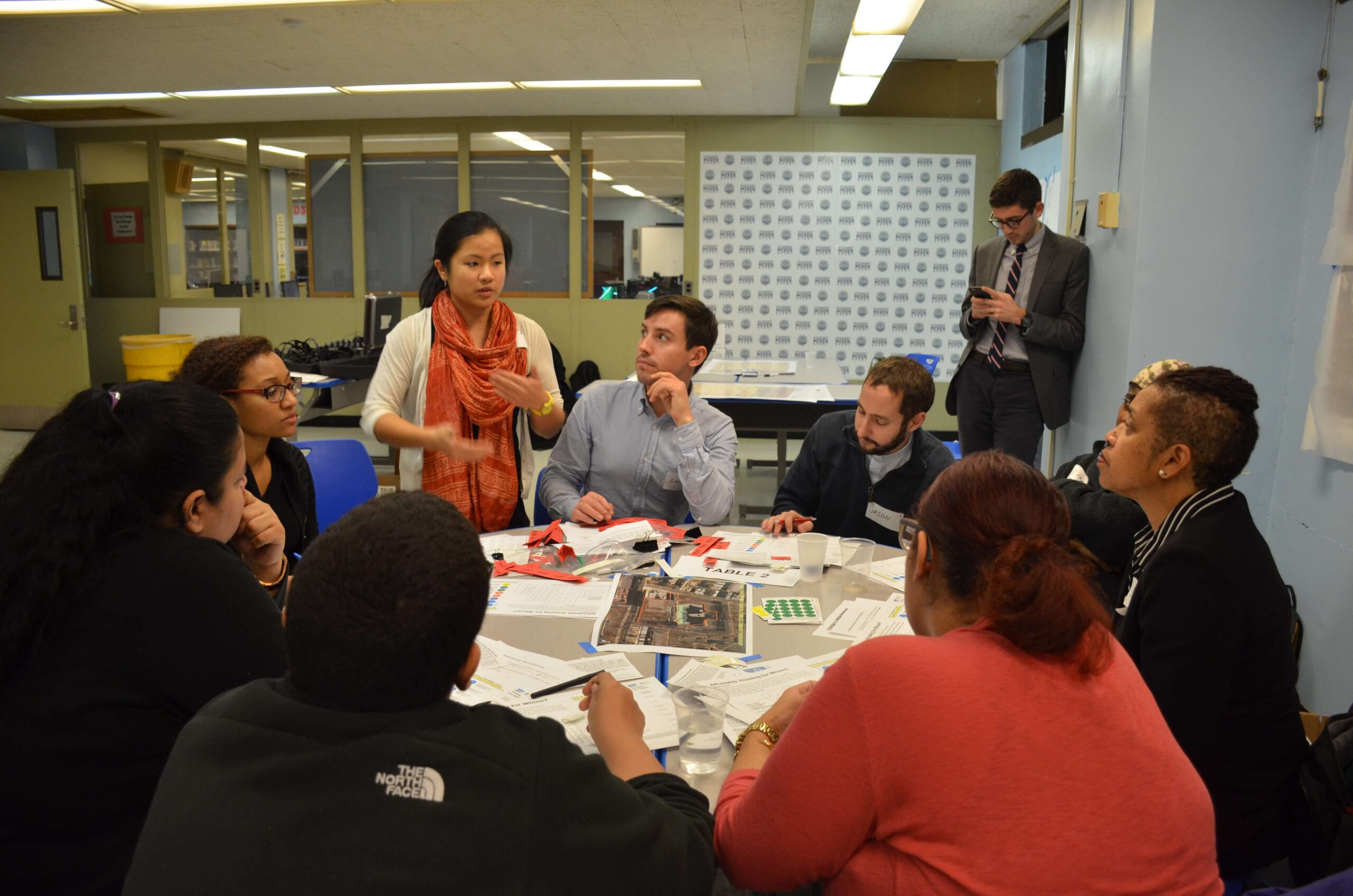 A group of people are gathered around a round table as they listen attentively to only person who is standing. There are various documents laid out on the table.
