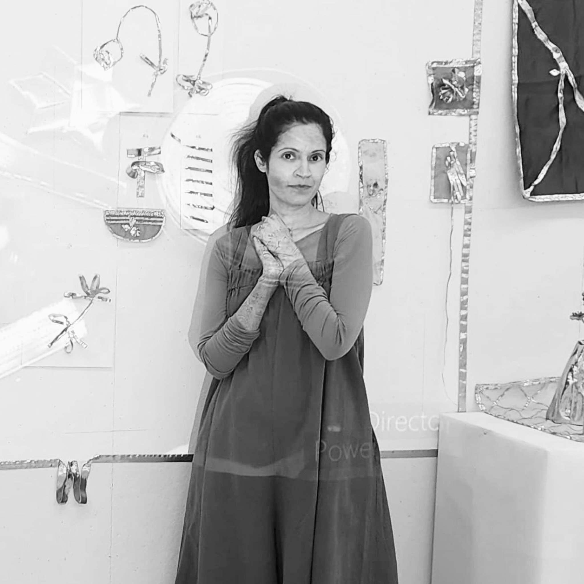 A woman stands in front of a wall-mounted mixed-media art installation. She wears a layered dress and long-sleeved top, with her hands clasped in front of her chest. The black and white image conveys a sense of artistic focus and presence.