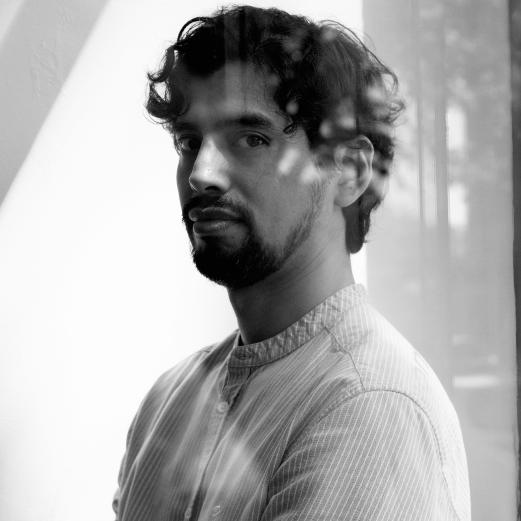 A man with curly hair and a trimmed beard stands indoors with natural light filtering through a nearby window, casting shadows on his face and shirt. He wears a striped button-down top and looks calmly at the camera. The portrait is in black and white.