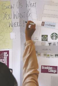A student is writing on a document pinned to the wall with a sharpie. It reads "Show us what you want & where!!"