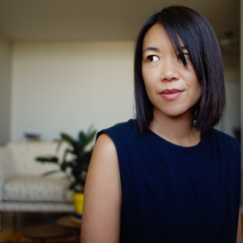 Minh-Ha T. Pham look to the side of the camera with a slight smile. In the background you can see a white couch with a glass table with a plant in front of it.