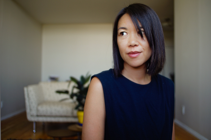 Minh-Ha T. Pham look to the side of the camera with a slight smile. In the background you can see a white couch with a glass table with a plant in front of it.