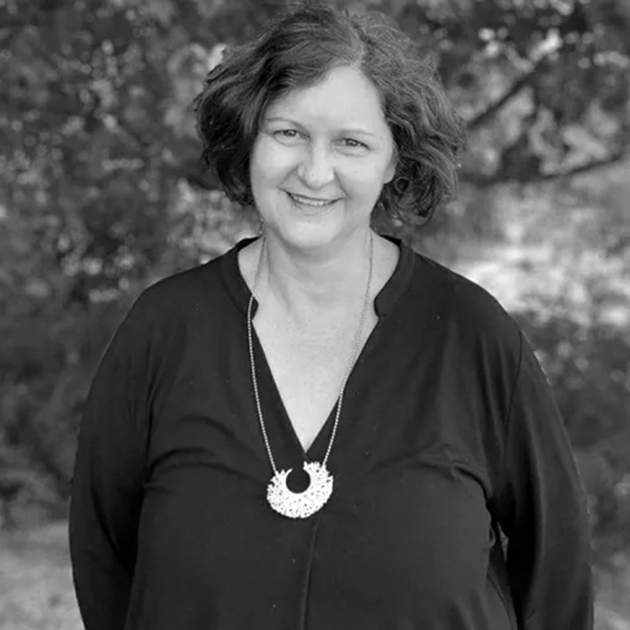 A middle-aged woman stands outdoors, smiling gently. She has short, wavy hair and wears a long necklace over a dark V-neck top. The background is blurred greenery, and the black and white image feels natural and warm.
