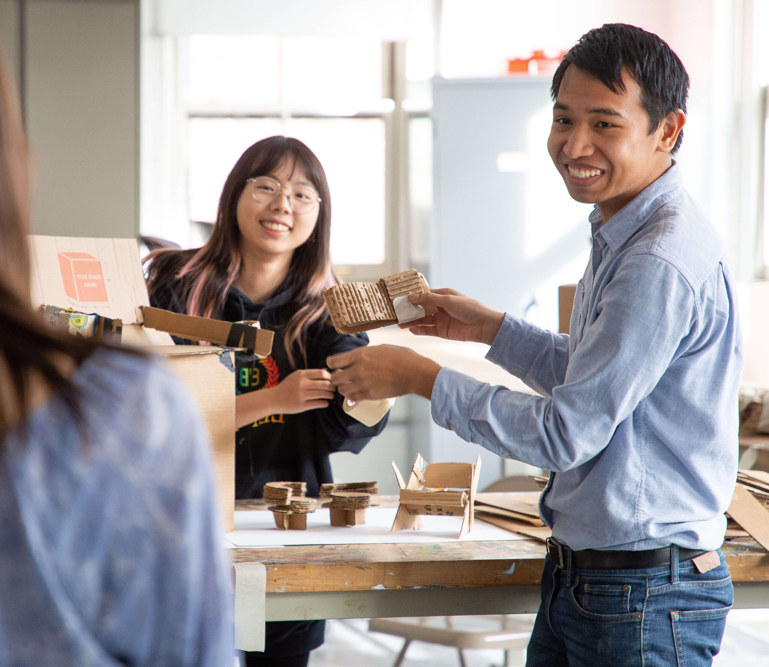 Two people show their works to a professor, whose back is partially in the picture. The student looks excited and proud of their work.