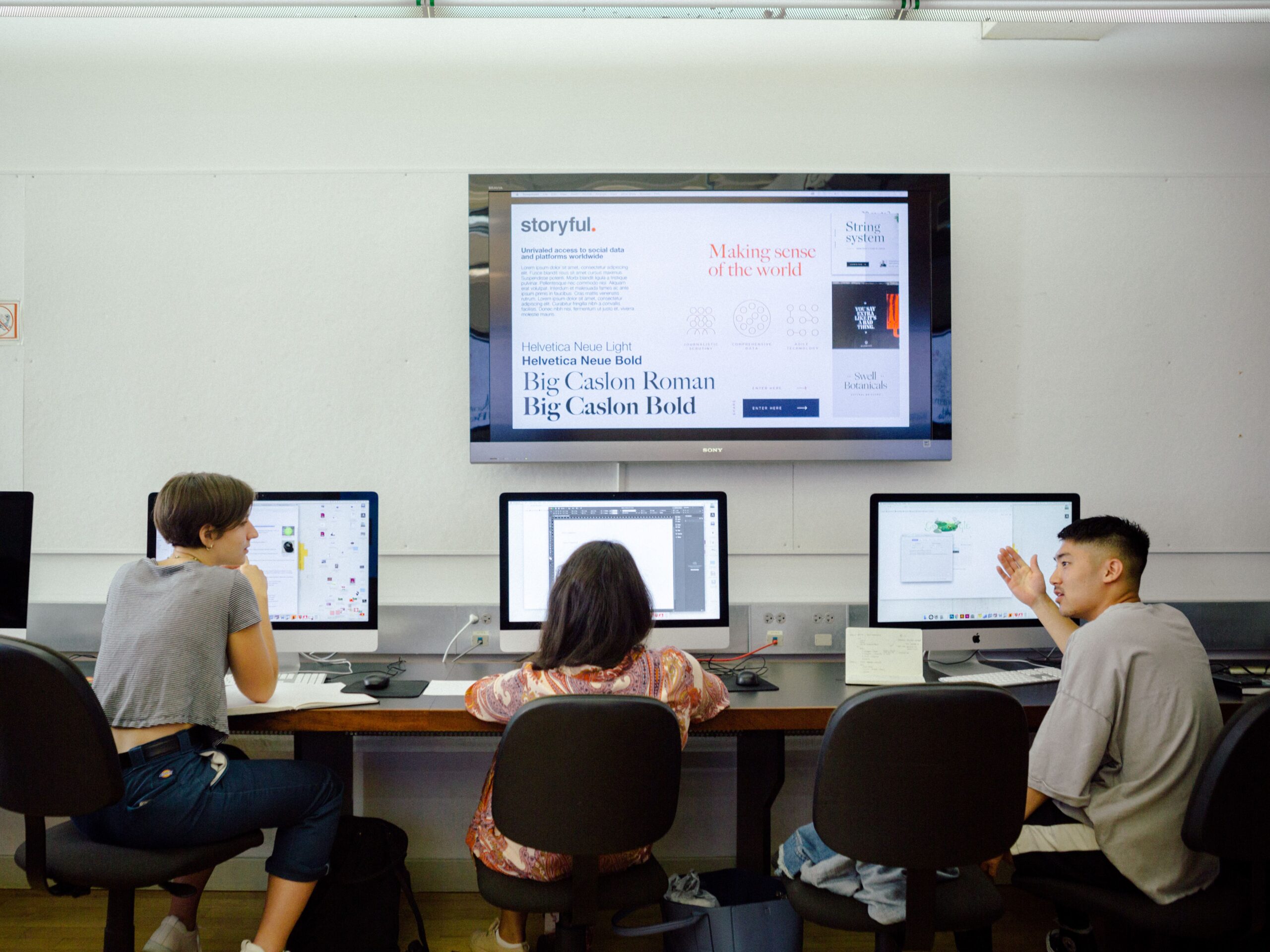 Three students sit in front their respective desktop computers. They are all facing away from the camera. The appear to be discussing amongst themselves. They are in a well lit room.