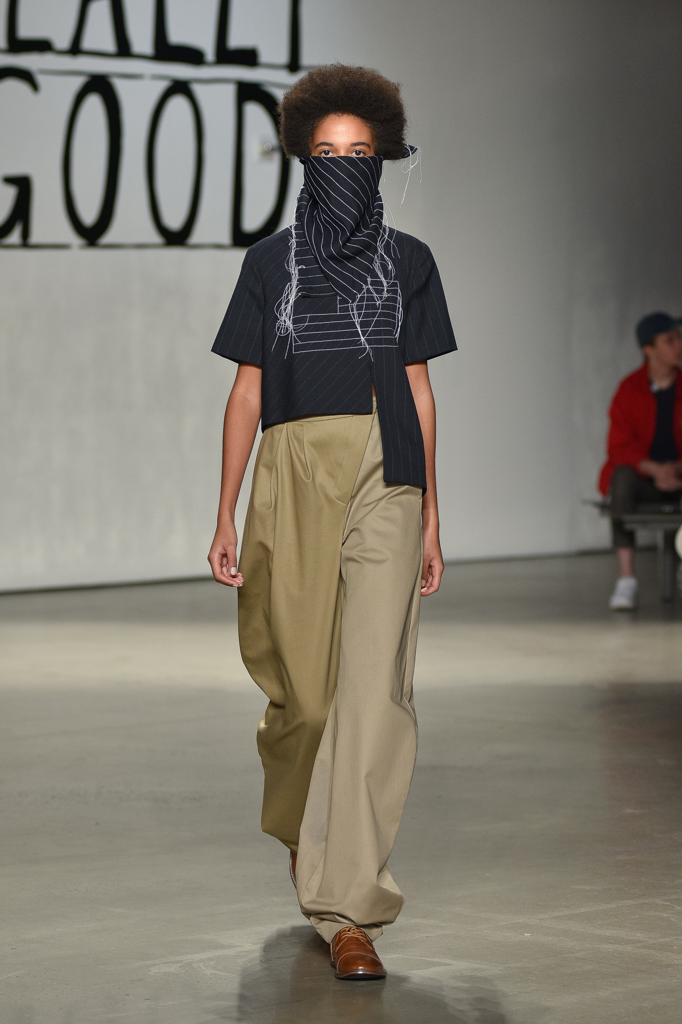 A model with a scarf covering her face walks down a runway. She is wearing a long beige trousers and a navy shirt.