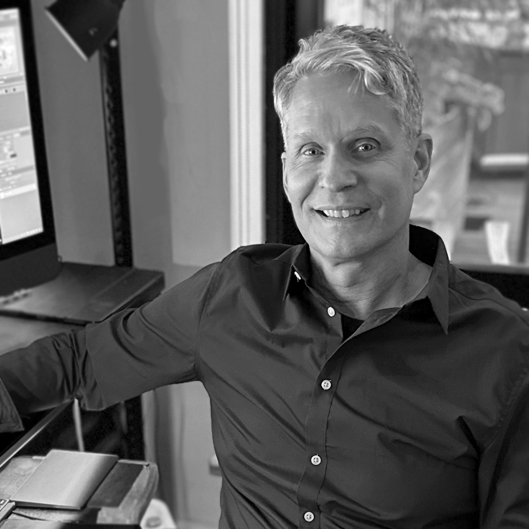 A man with short silver hair smiles at the camera while seated at a desk with a large monitor. He wears a dark button-up shirt. The setting appears to be a home office or creative workspace. The photo is black and white and inviting.