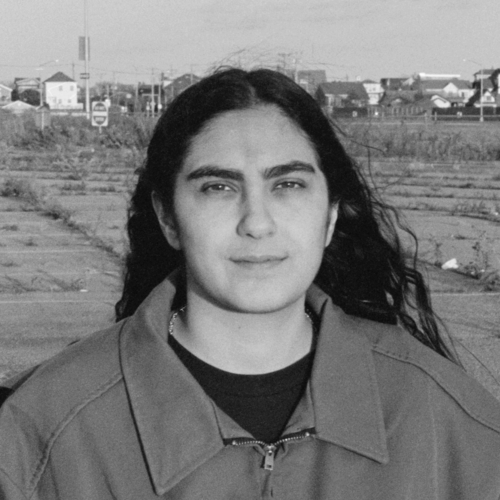 A woman with long dark hair stands in an open outdoor space, looking calmly at the camera. She wears a zip-up jacket, and behind her is a vast, empty lot with suburban houses and signage in the distance. The photo is in black and white.