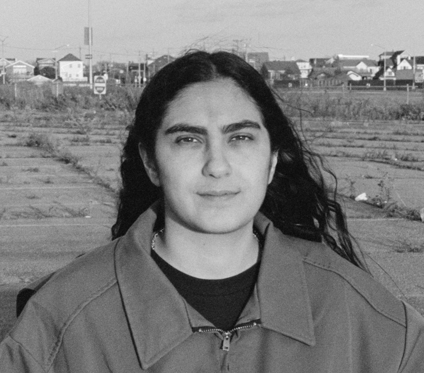 A woman with long dark hair stands in an open outdoor space, looking calmly at the camera. She wears a zip-up jacket, and behind her is a vast, empty lot with suburban houses and signage in the distance. The photo is in black and white.