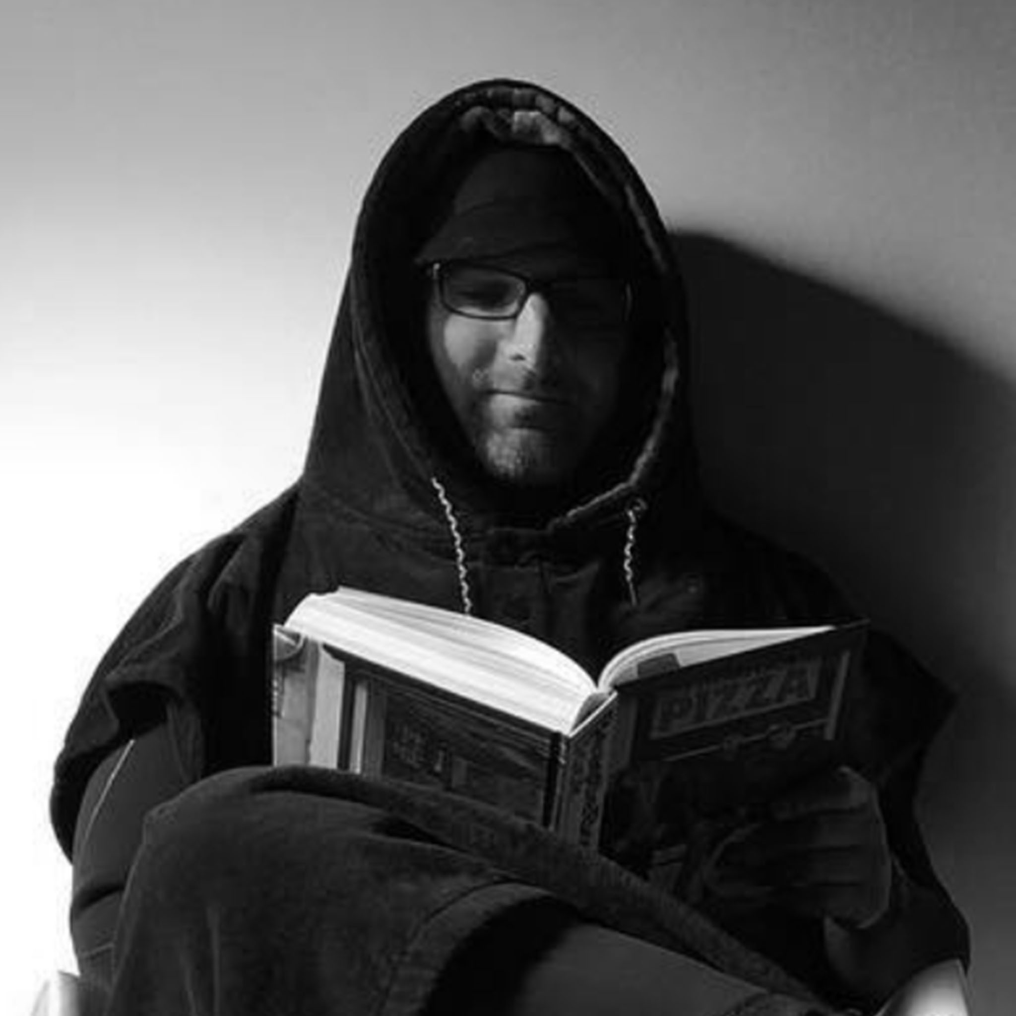 A person sits comfortably, wearing a hooded blanket or robe and glasses, reading a book titled “PIZZA.” The image is black and white, with a strong shadow cast on the wall, creating a cozy, contemplative atmosphere.