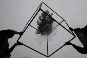 Low-angle view of multiple people holding the corners of a square wooden frame above their heads against an overcast sky. Suspended within the frame is a semi-transparent sculptural form made of dark, cloud-like mesh and lighter geometric elements, resembling an abstract mass or digital terrain. The people are visible only as partial silhouettes—arms, hands, and shoulders—emphasizing the structure and artwork rather than individual identities.