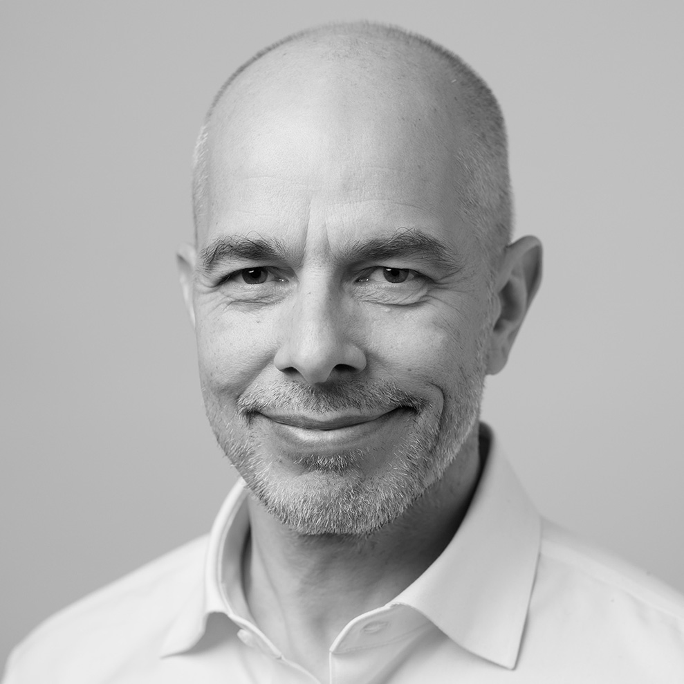 A bald man with a close-cropped gray beard smiles slightly at the camera. He is wearing a white button-up shirt and stands in front of a light gray background. The image is in black and white.
