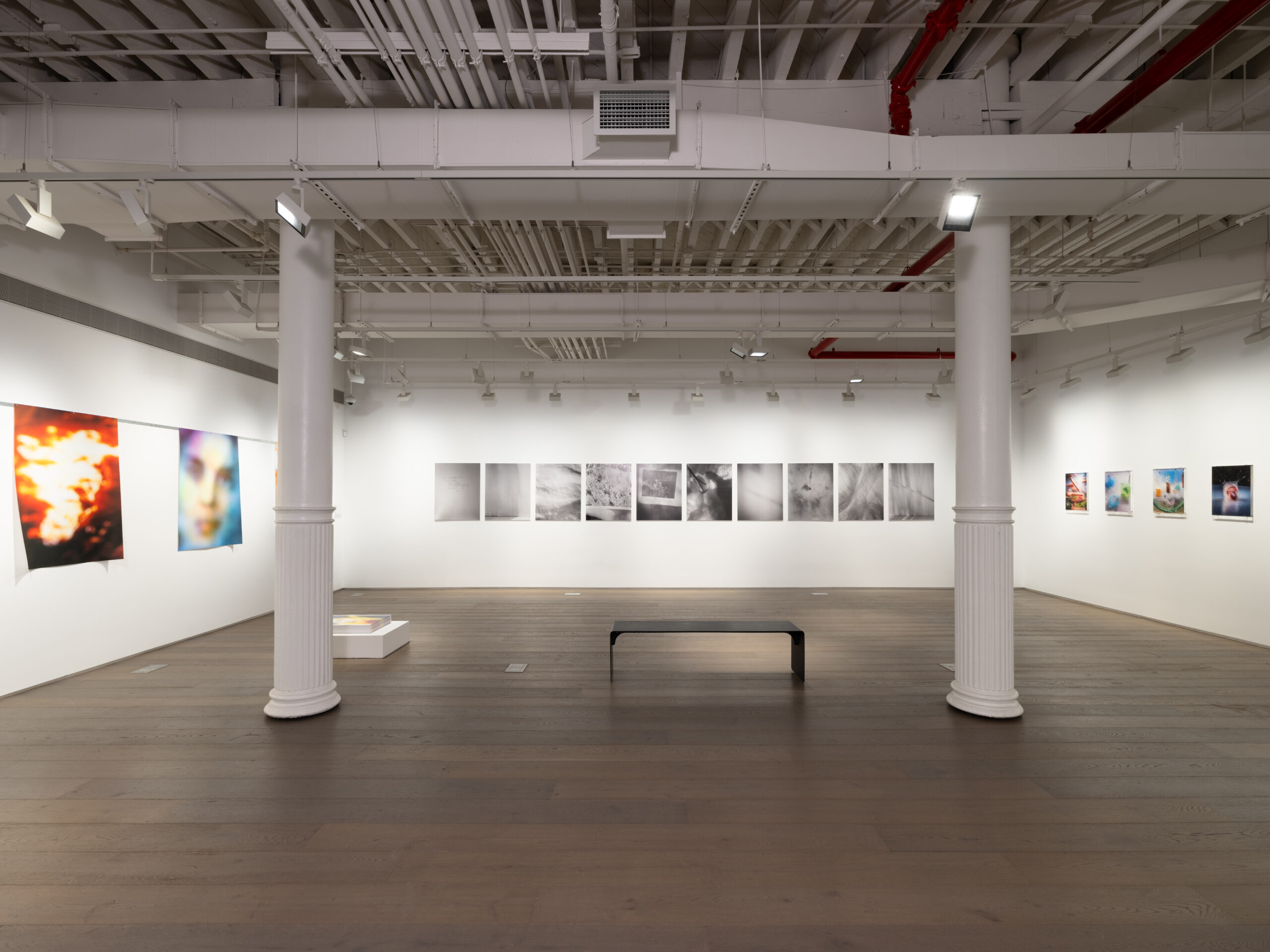 A contemporary art gallery with white walls, an open ceiling exposing white-painted pipes, and wooden flooring. The room is illuminated by ceiling-mounted spotlights. Various photographic artworks are displayed on the walls, with images ranging from abstract and blurred portraits to black-and-white conceptual photography. A black bench is positioned in the center of the room, and a white pedestal holding a book or artwork is placed towards the left side. The gallery has a minimalist aesthetic with two white columns supporting the ceiling.