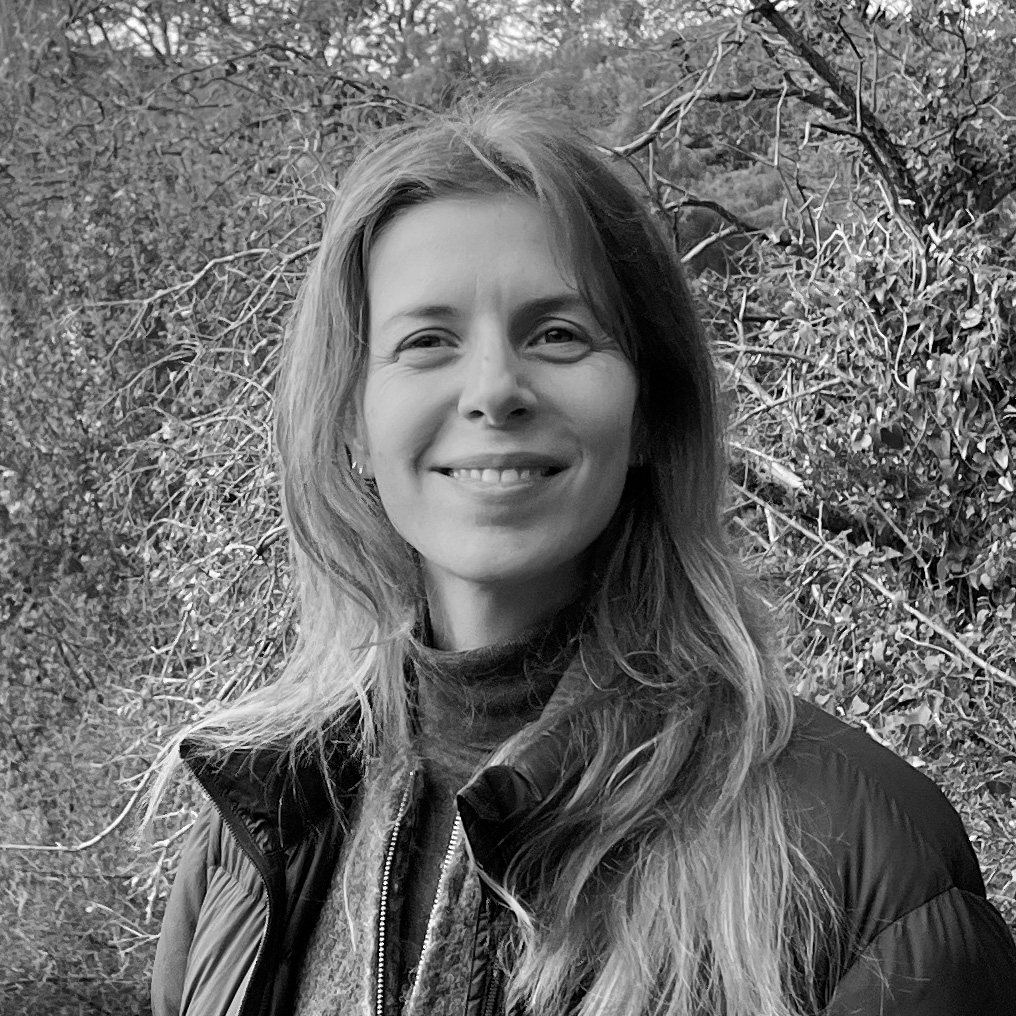 A woman with long wavy hair stands outdoors, smiling at the camera. She wears a turtleneck sweater and a puffy jacket, with bare tree branches forming the background. The photo is black and white, conveying a sense of calm and nature.