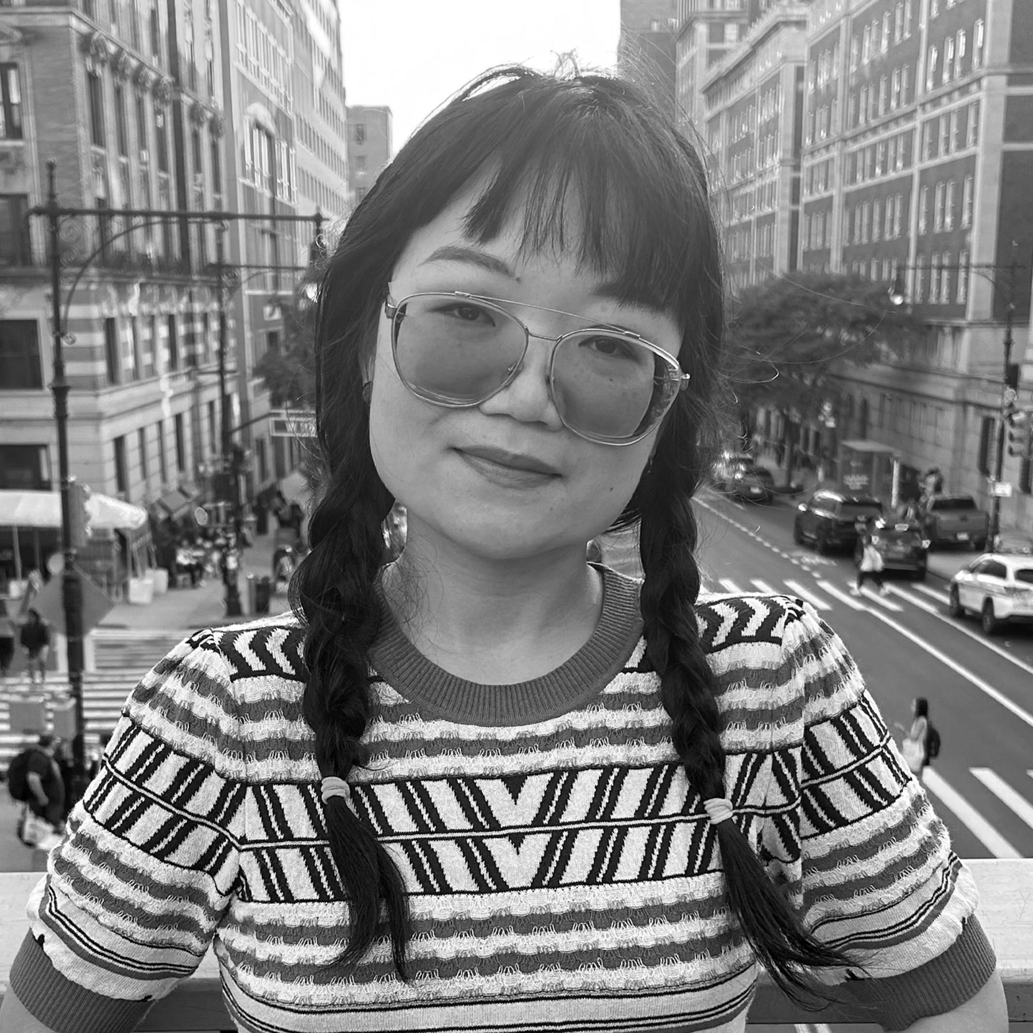 A young woman with glasses and braided pigtails stands on a balcony overlooking a city street. She wears a patterned shirt and smiles slightly. The photo is black and white and captures a candid urban moment.