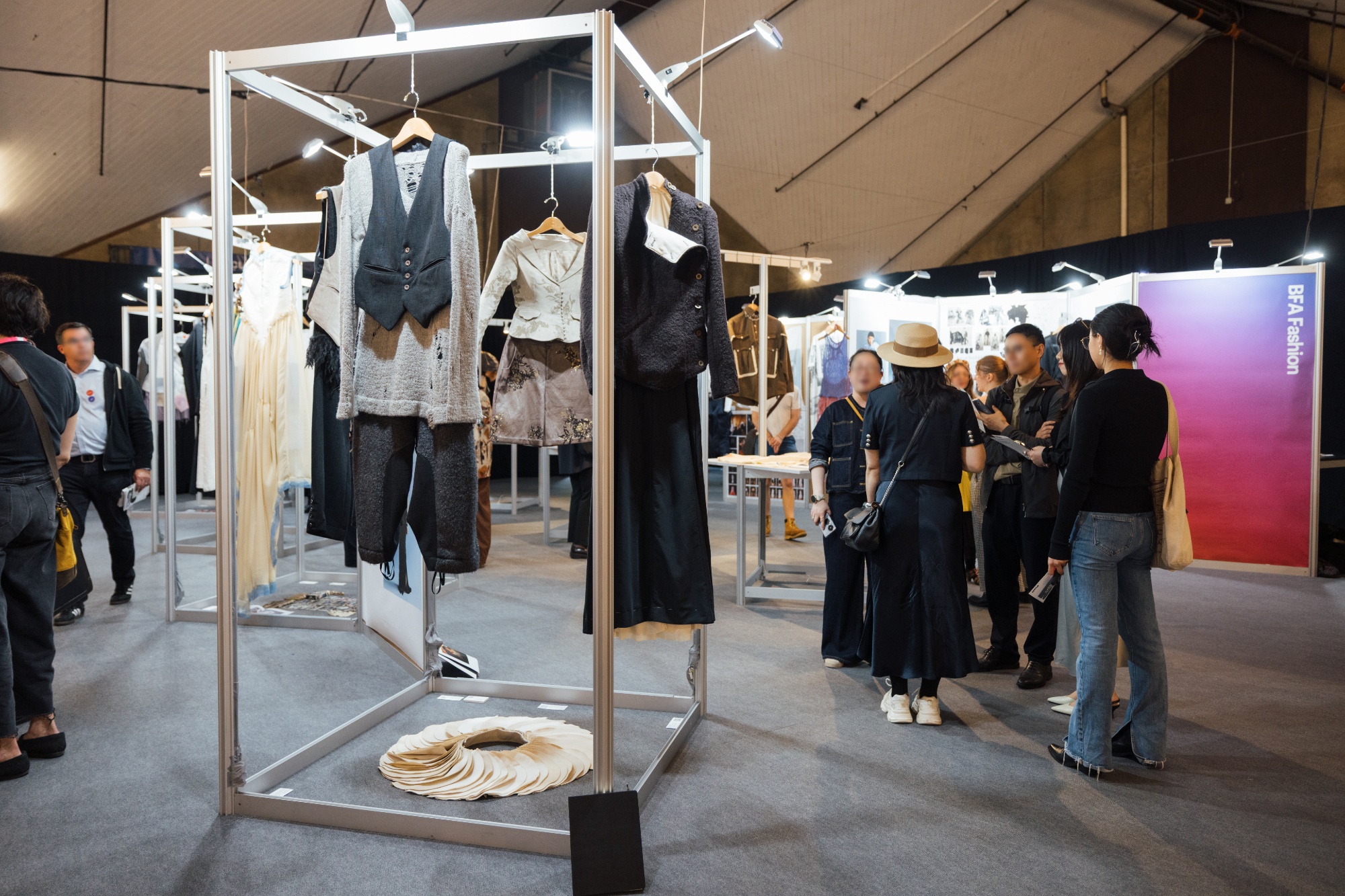 A fashion exhibition scene showing garments displayed on minimalist metal racks under gallery lighting, with visitors standing nearby and examining the clothing in a large open hall.
