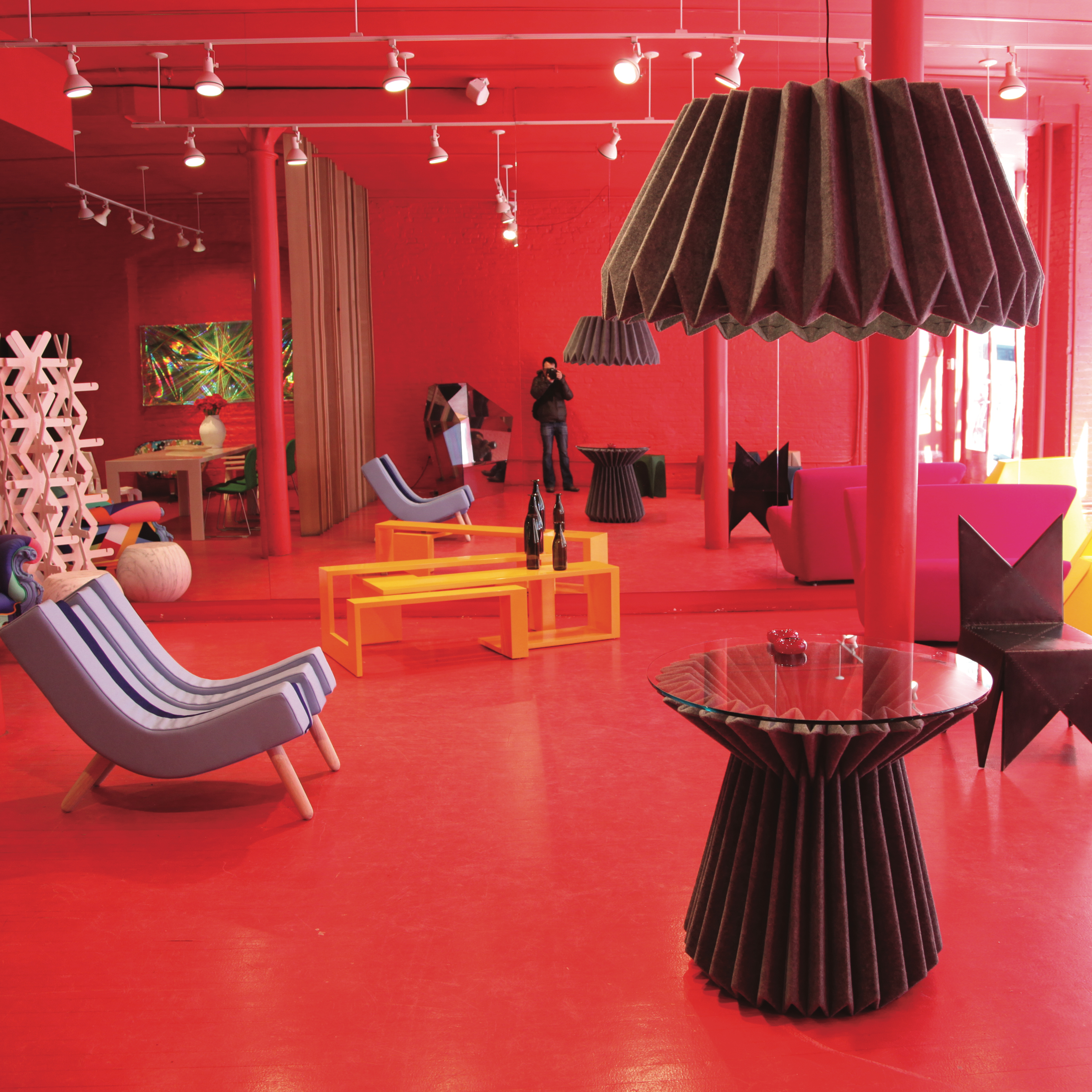 A red colored room with an assortment of different furniture pieces and decorations. The decorations are made of many colors, including yellow, maroon, purple, green, black, pink and more. There is a large mirror at the back of the room where the reflection of the person taking the picture can be seen.