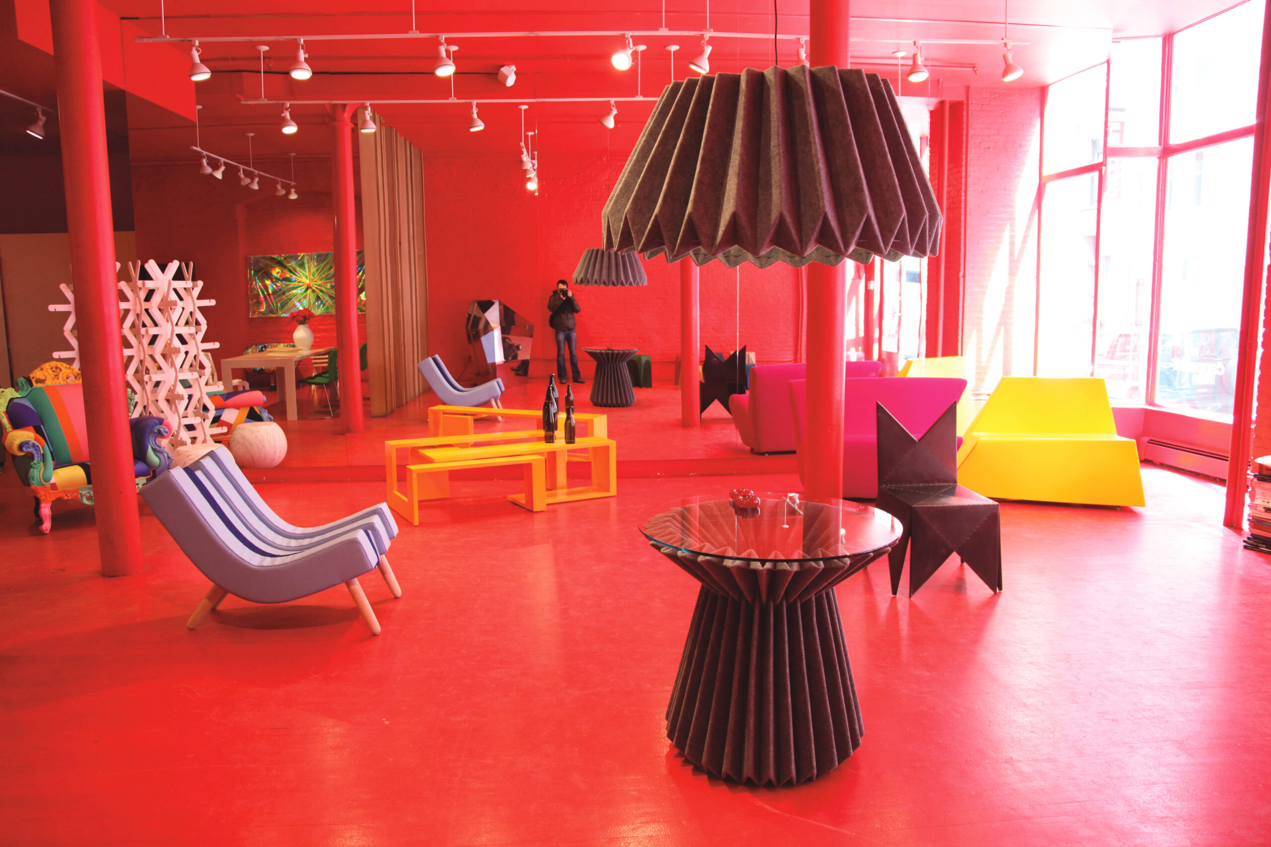 A red colored room with an assortment of different furniture pieces and decorations. The decorations are made of many colors, including yellow, maroon, purple, green, black, pink and more. There is a large mirror at the back of the room where the reflection of the person taking the picture can be seen.
