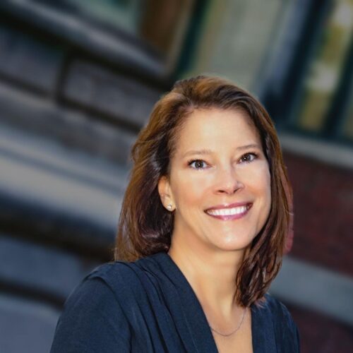 Professional headshot of a woman smiling outdoors, wearing a dark navy dress with a pleated neckline. The background is softly blurred, showing a brick building and architectural details.