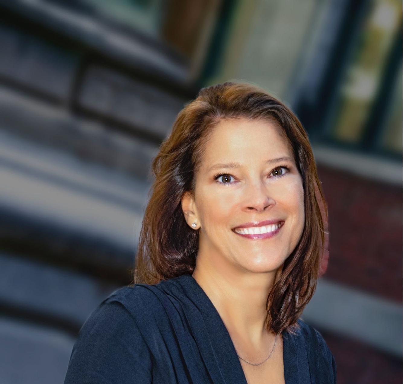 Professional headshot of a woman smiling outdoors, wearing a dark navy dress with a pleated neckline. The background is softly blurred, showing a brick building and architectural details.