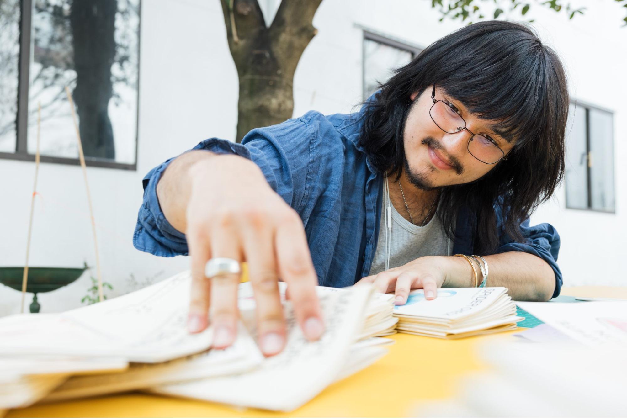 Close-up of a person seated outdoors at a yellow table, turning the pages of a small booklet or pamphlet. The individual has shoulder-length dark hair, wears glasses and a blue shirt, and is focused on the printed material in their hands. A tree trunk and a white building facade appear softly out of focus in the background.