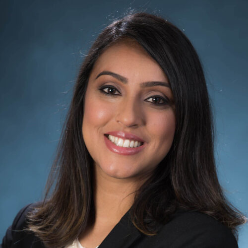 Professional headshot of a woman smiling confidently, wearing a black blazer and white blouse against a blue gradient studio background.