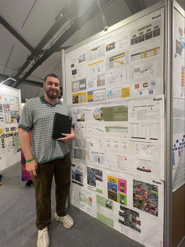 A person stands smiling next to a large presentation board at an exhibition. The board displays architectural and project management materials, including charts, site plans, safety diagrams, and logistics schedules, with the Pratt Institute logo visible in multiple places. The person holds a black folder and appears to be presenting their capstone project in a well-lit indoor academic setting.