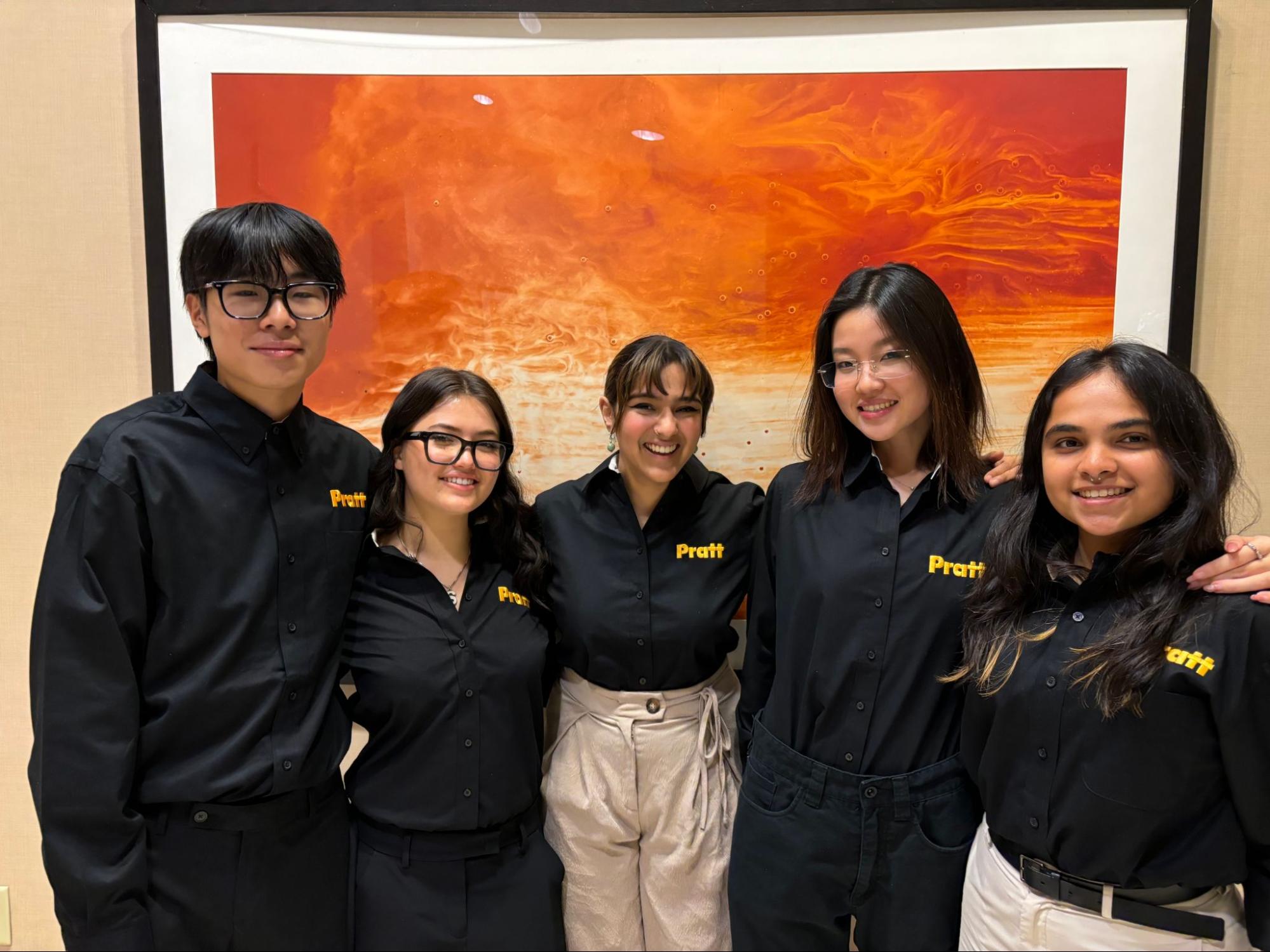 Five students wearing black shirts embroidered with the Pratt logo stand smiling in front of a large orange abstract artwork, posing for a group photo.