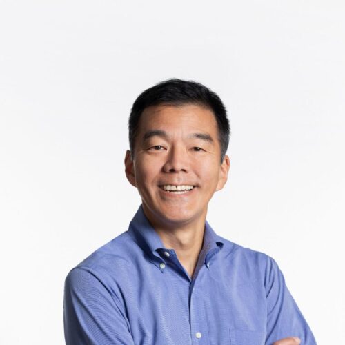 Portrait of a man smiling with arms crossed, wearing a blue button-down shirt and black jeans, standing against a plain white background.