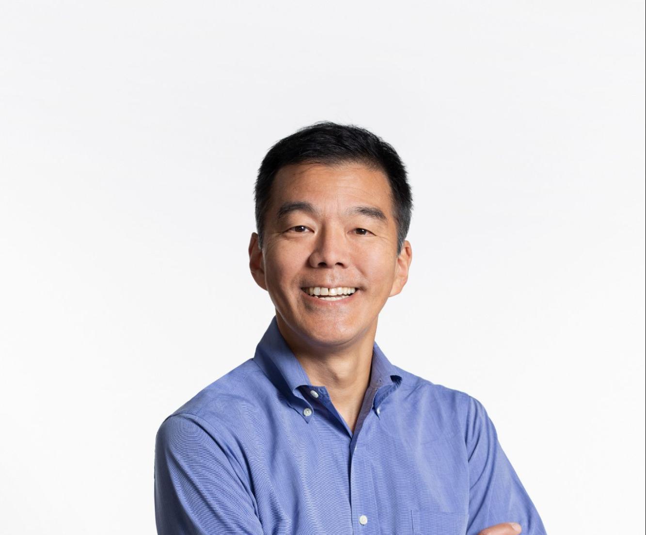 Portrait of a man smiling with arms crossed, wearing a blue button-down shirt and black jeans, standing against a plain white background.