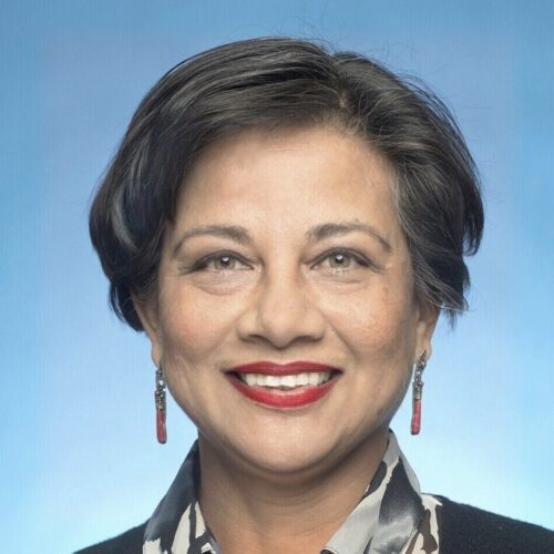 Headshot of a woman smiling warmly against a blue background, wearing a patterned blouse, black cardigan, and red earrings.