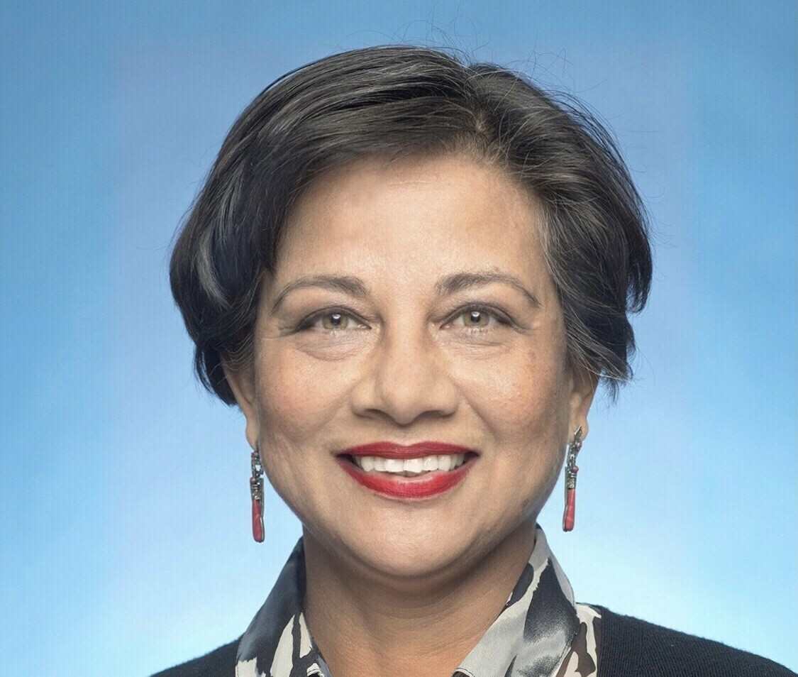 Headshot of a woman smiling warmly against a blue background, wearing a patterned blouse, black cardigan, and red earrings.