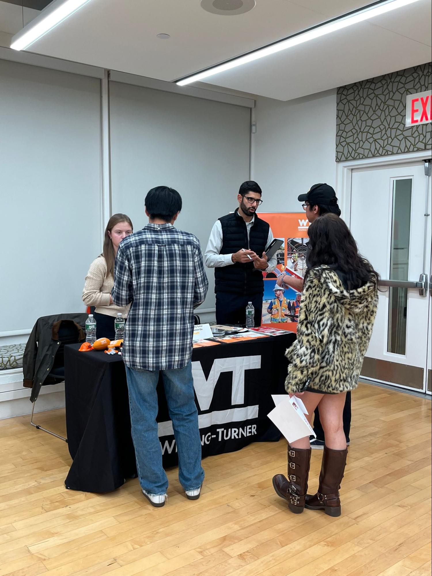 Students speak with representatives at a W.T. (Whiting-Turner) information table during a campus career or networking event. The table is covered with brochures and branded materials, and the interaction takes place in a well-lit room with light wood flooring and neutral walls.