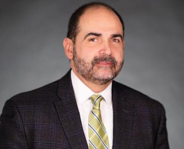 Portrait of a man wearing a dark checkered suit jacket, light shirt, and green striped tie, posing against a gray studio background.