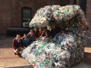 group of students pantomiming surfing, in a sculpture made of recyclable refuse consisting of bottles, packaging, etc