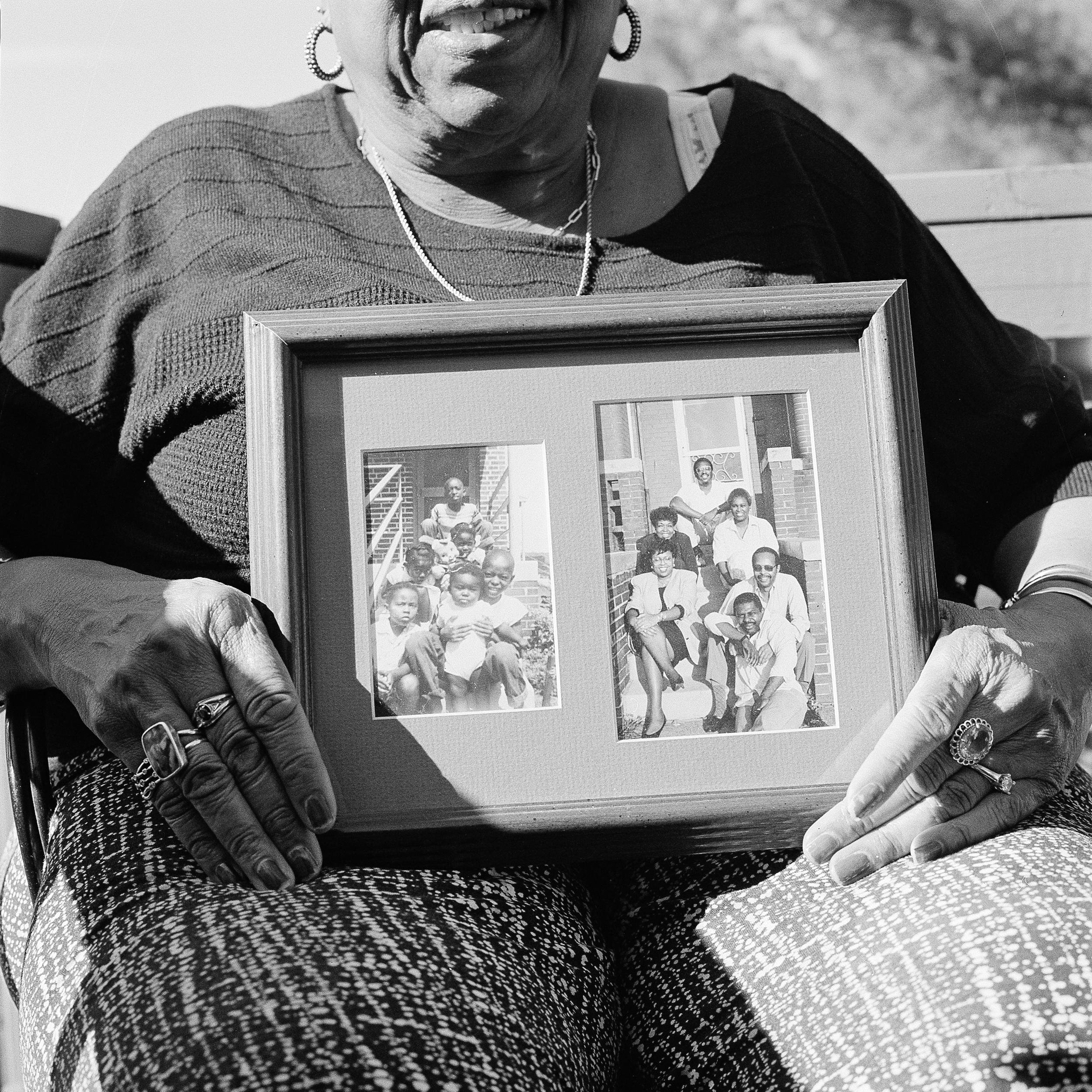 A black and white photograph of a woman seated outdoors, holding a framed photograph with two images. The image on the left shows a group of children sitting together, while the right side features adults posing in front of a building. The woman wears a black top and patterned pants, adorned with several rings. Her hands prominently display the frame, and she smiles gently while looking at it.