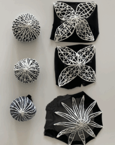 A collection of black fabric panels and spherical forms with white 3D-printed lattice structures applied on top. The designs resemble floral or starburst patterns, including four-petal and radial arrangements. The contrast of white lattice against black fabric highlights the sculptural detail.