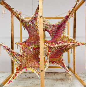 A woven installation made of multicolored yarn (red, yellow, blue, cream) stretched into star-like geometric forms within a wooden cubic frame. The handwoven netting creates interconnected, organic shapes, resembling textile-based architectural or sculptural exploration.