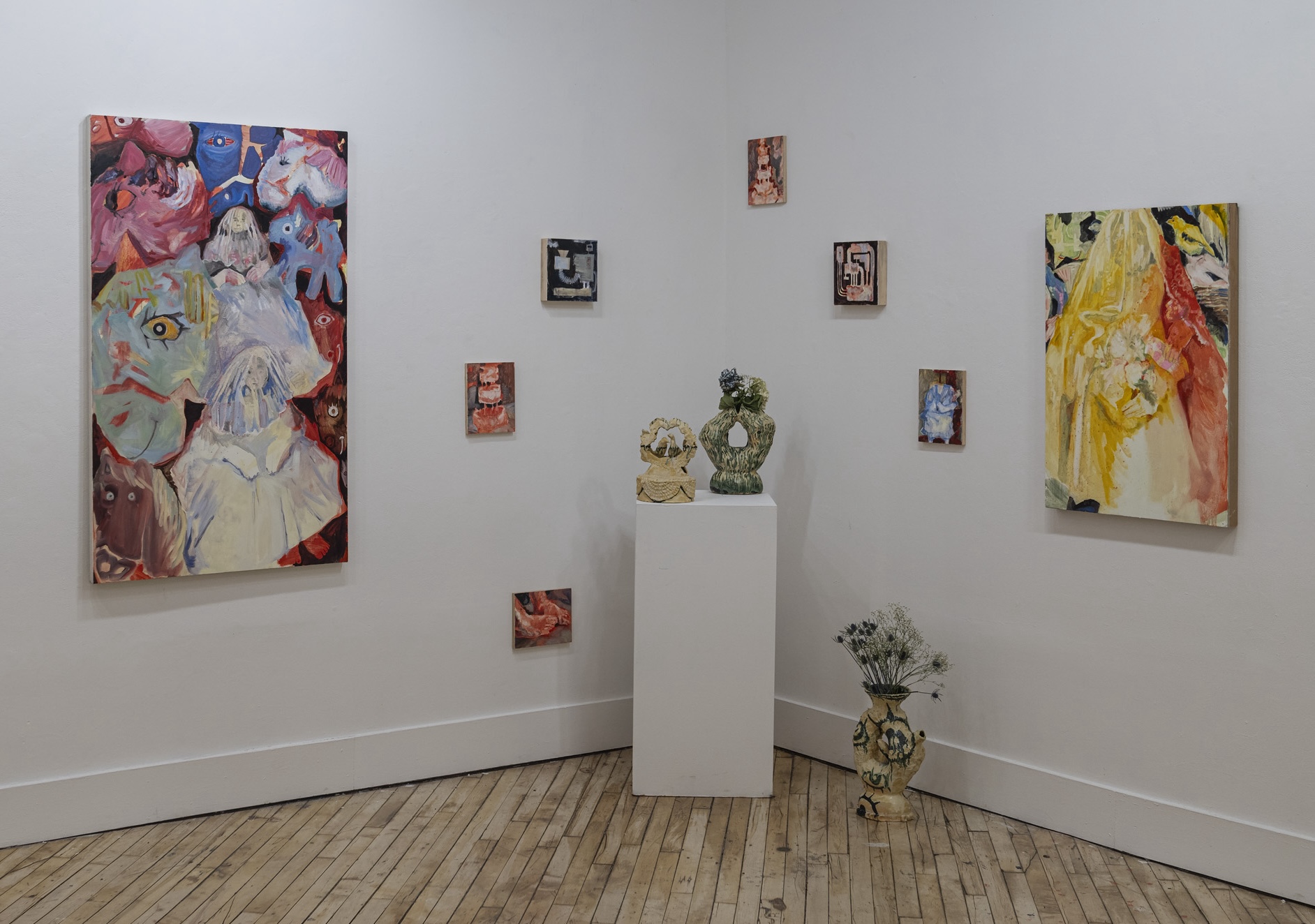 An art gallery corner display featuring a collection of paintings and ceramic sculptures. The walls are adorned with several expressive, colorful paintings, including two large canvases on either side of the corner and several smaller pieces arranged between them. The paintings depict abstract and surreal figures and forms, with vibrant and contrasting colors. In the corner, a white pedestal holds two intricately designed ceramic sculptures, and a ceramic vase with a floral arrangement is placed on the floor nearby. The setting is an intimate gallery space with wooden floors, emphasizing the creative and artistic atmosphere of the exhibition.