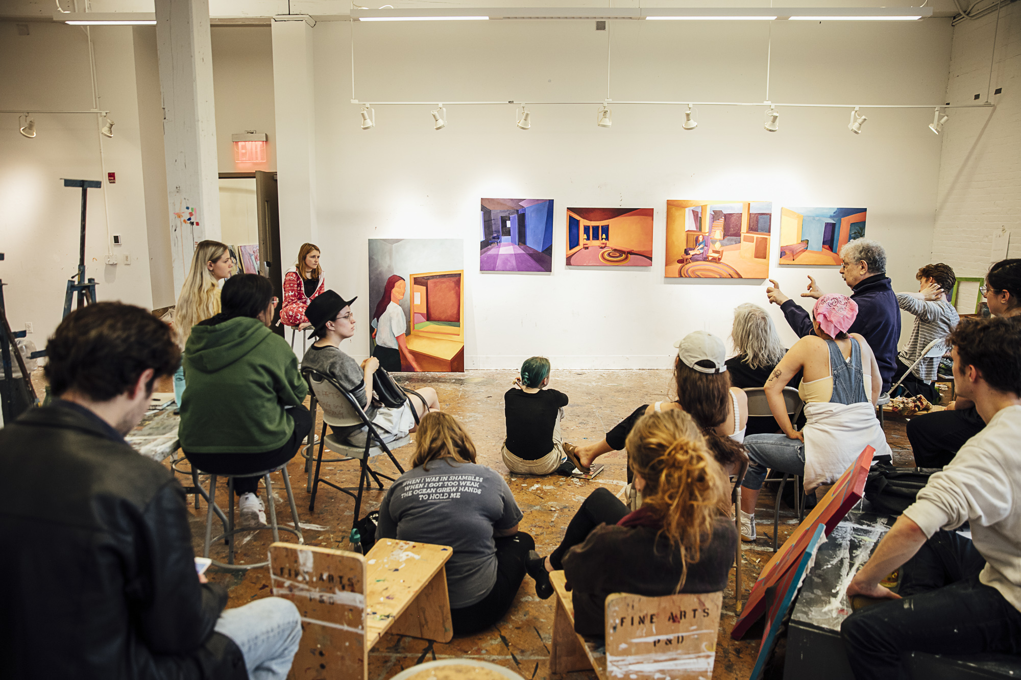 Students sit in a painting studio as a professor lectures on 5 paintings shown at the front of the classroom.