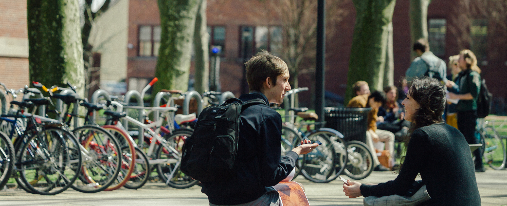 Tow students chat while sitting in front of a bike rack packed with bikes