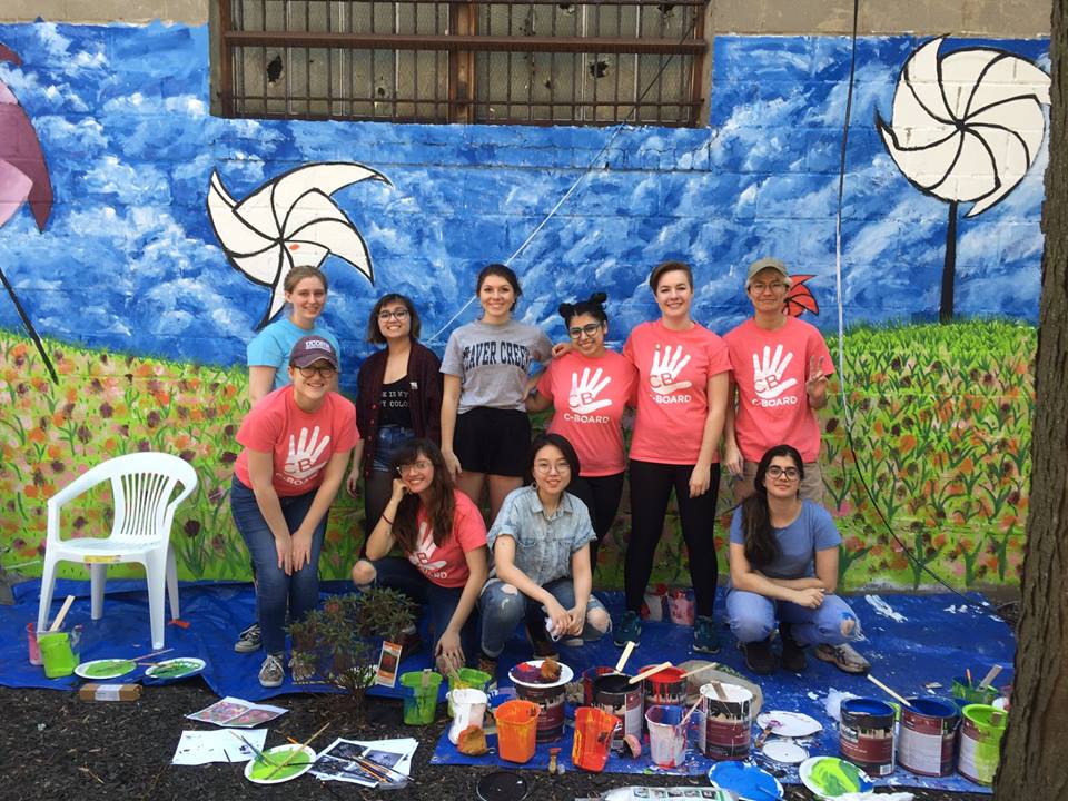 A group of students pose in front of a mural depicting a green fields with flowers and a blue sky with windmills scattered throughout. The students are surrounded by painting equipment.