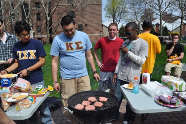 Fraternity members are pictured grilling hamburgers in a field as they socialize.