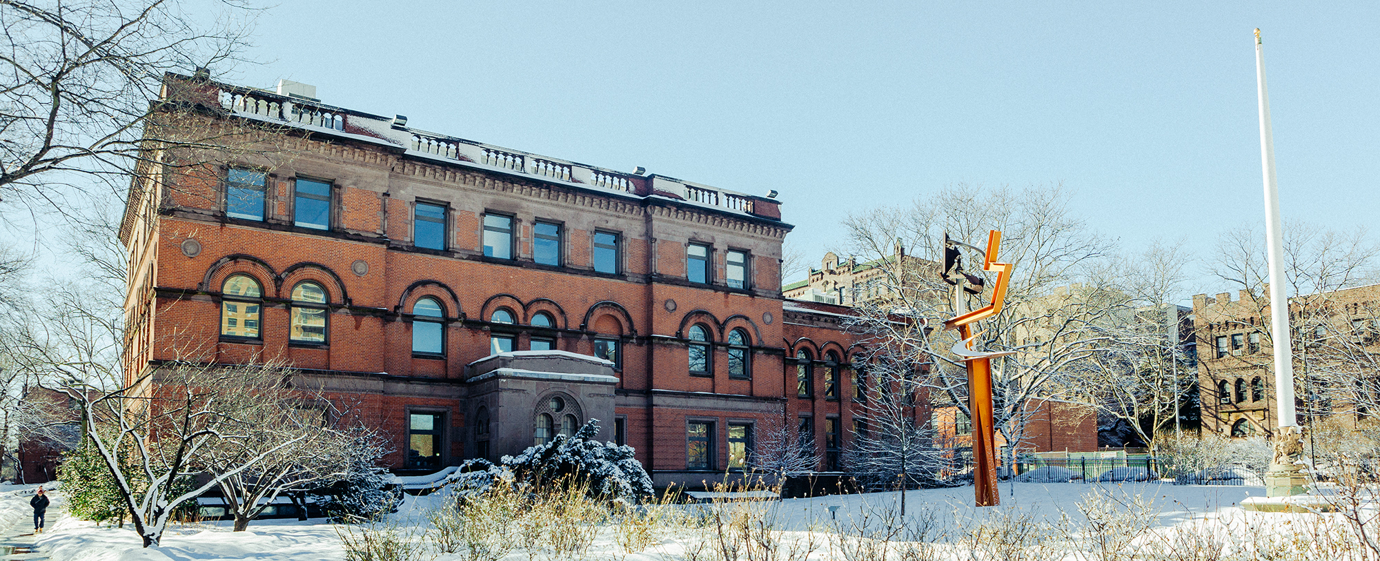 Photo of the a red brick building with a sculpture and yard in front of it. Everything is covered by a thing layer of snow and it is very bright.